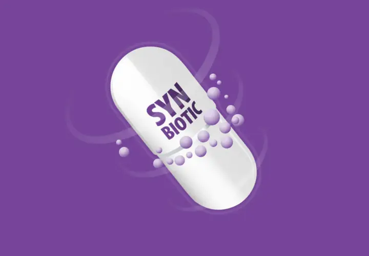 Biała kapsułka z napisem "SYN BIOTIC" na fioletowym tle. Drobne kuleczki unoszą się wokół. Dowiedz się, synbiotyk co to.